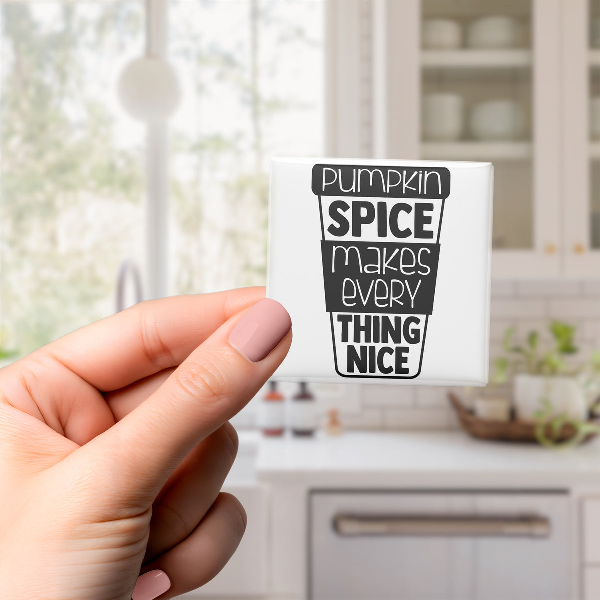 A hand holding a white magnet with a black text design that says "Pumpkin Spice Makes Every Thing Nice".