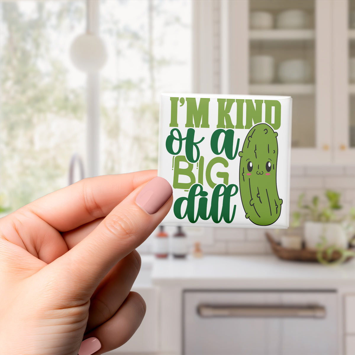 A hand holding a sticker with a cartoon image of a pickle and the text "I'm kind of a big dill" against a blurred background of a kitchen.