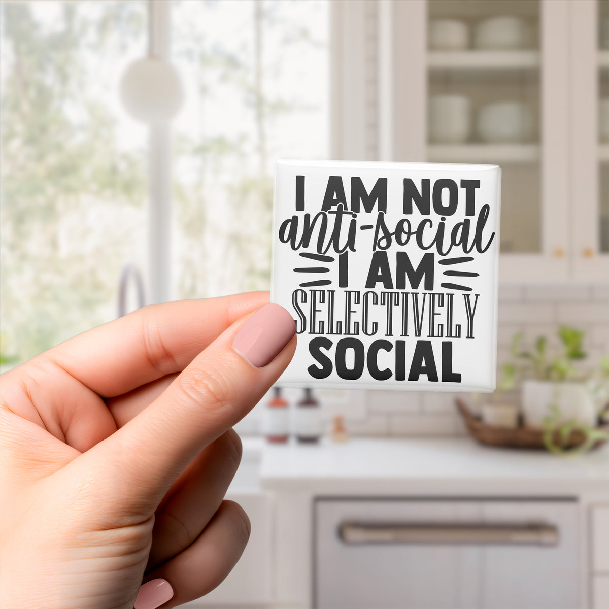 A person's hand holding a sticker with the text "I am not anti-social I am selectively social" against a blurred background of a kitchen.