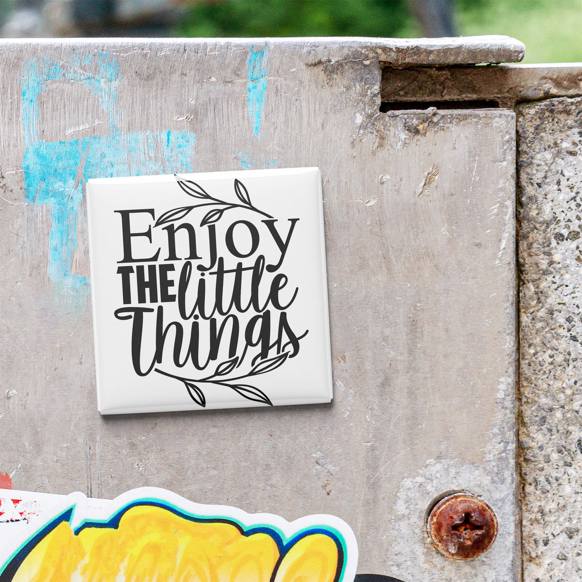 a white square magnet with the text "Enjoy the little things" written on it, placed on a concrete surface.