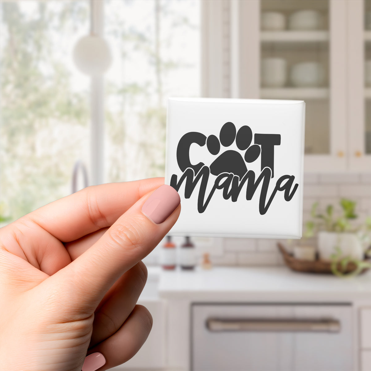 A hand holding a square magnet with the text "CAT MAMA" on it.