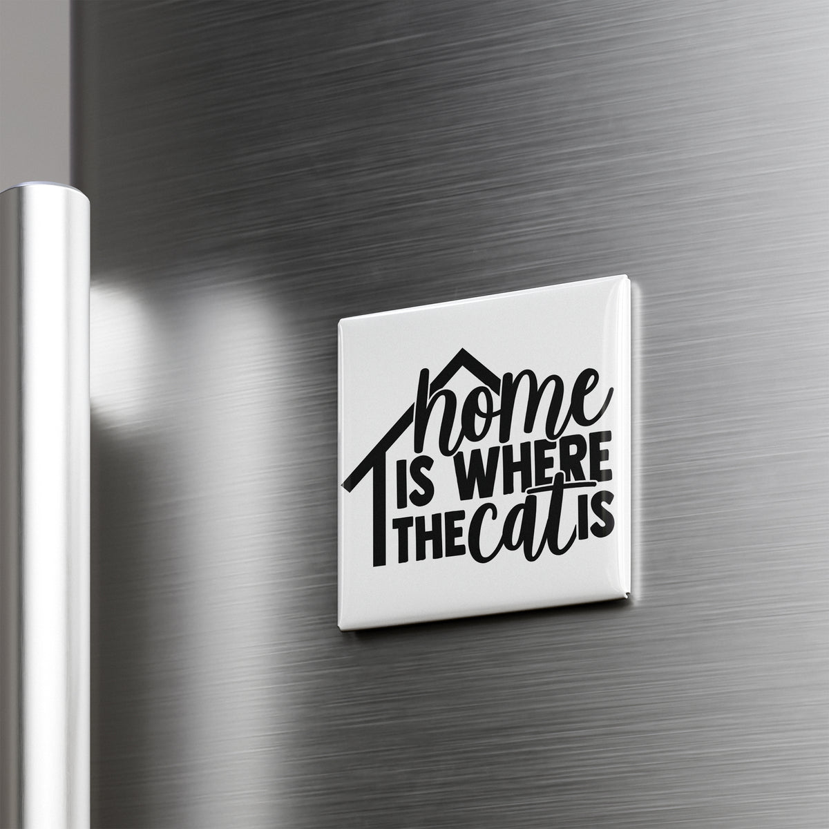 A white square magnet with the text "home is where the cat is" printed on it, mounted on a metallic surface.