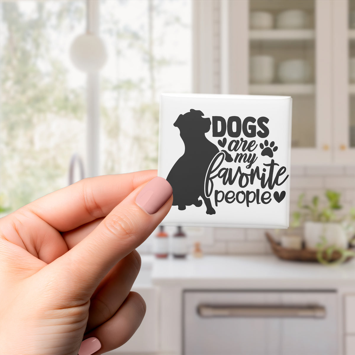 A hand holding a small square magnet with a black silhouette of a dog and the text "Dogs are my favorite people" printed on it.