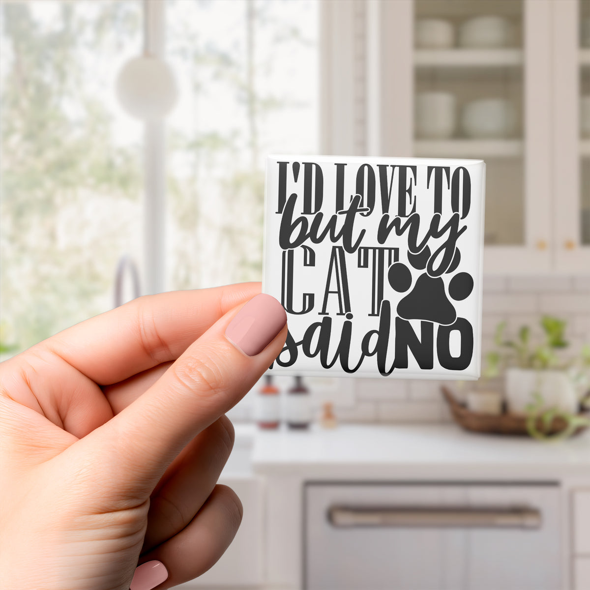 A hand holding a sticker with the text "I'd love to buy my cat said no" against a blurred background of a kitchen counter and cabinets.