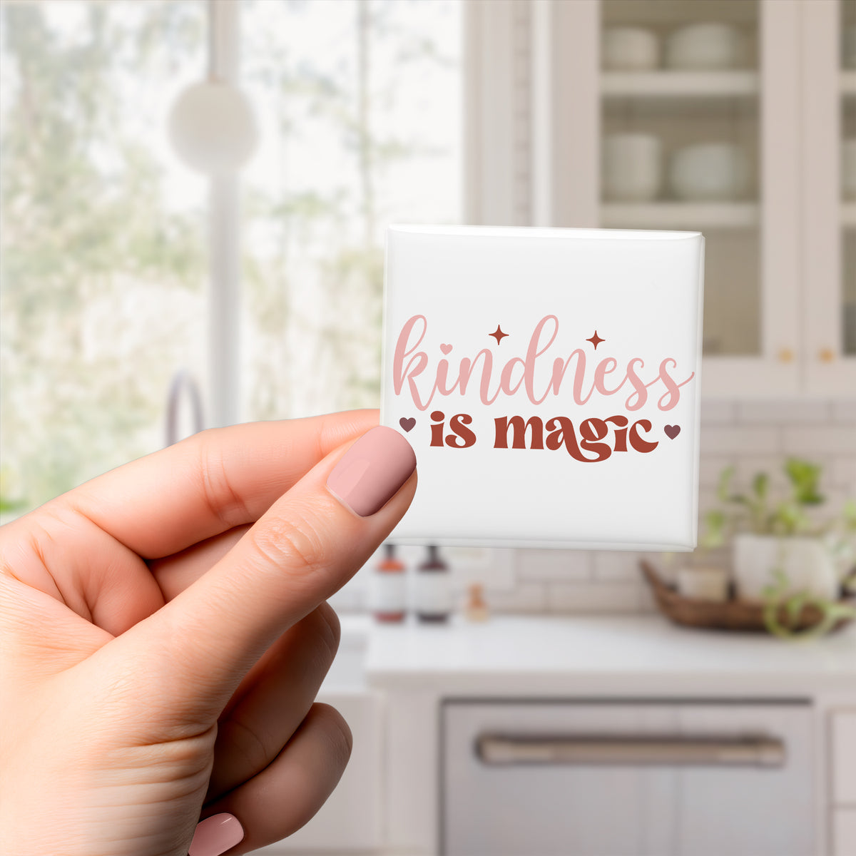 A hand holding a small square magnet with the text "kindness is magic" written on it.