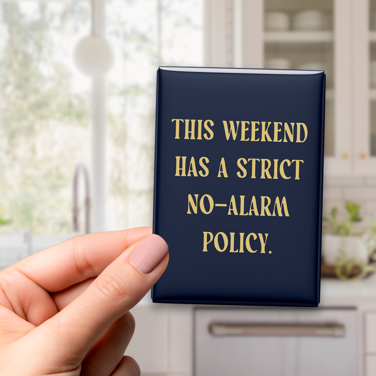 Hand holding the 2.5 × 3.5-inch “This weekend has a strict no-alarm policy.” magnet in a kitchen