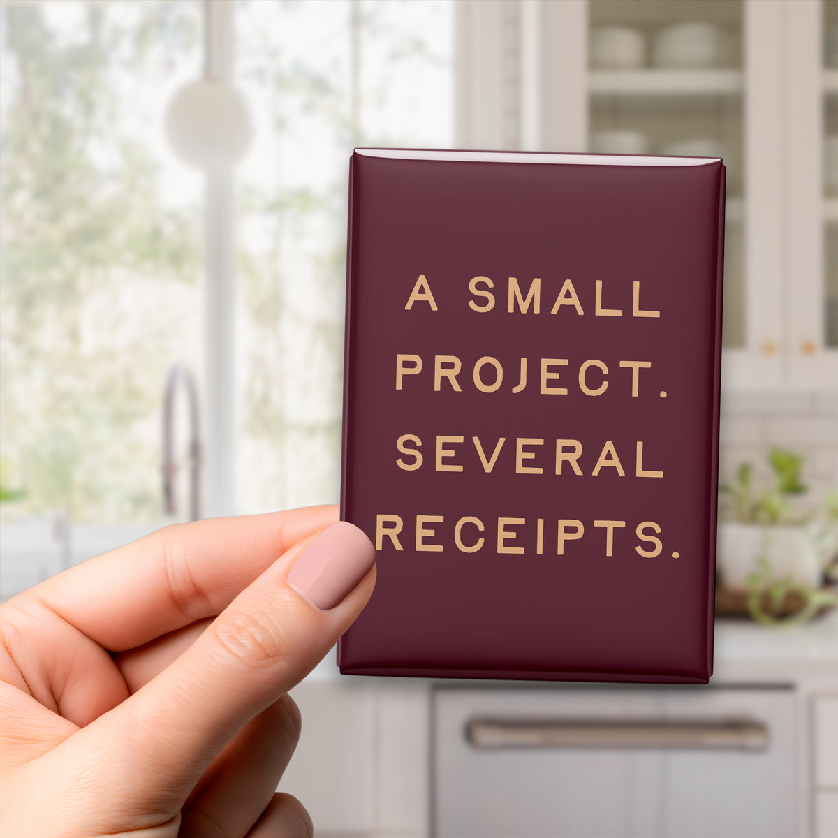 Hand holding the 2.5 × 3.5-inch “A small project. Several receipts.” magnet in a kitchen