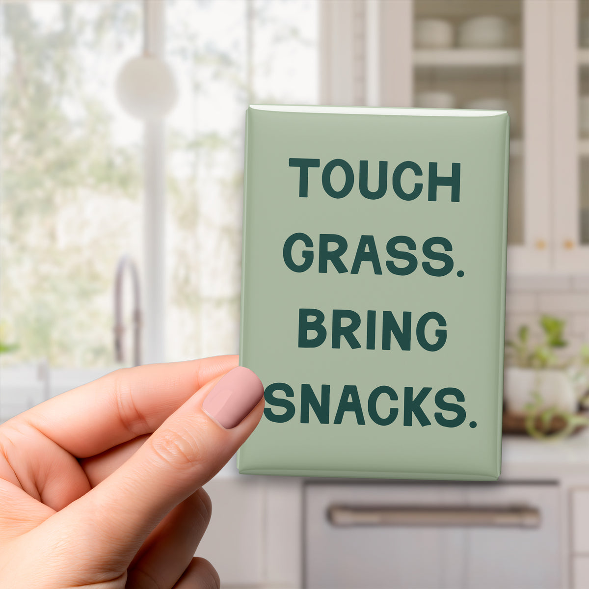 Hand holding the 2.5 × 3.5-inch “Touch grass. Bring snacks.” magnet in a kitchen