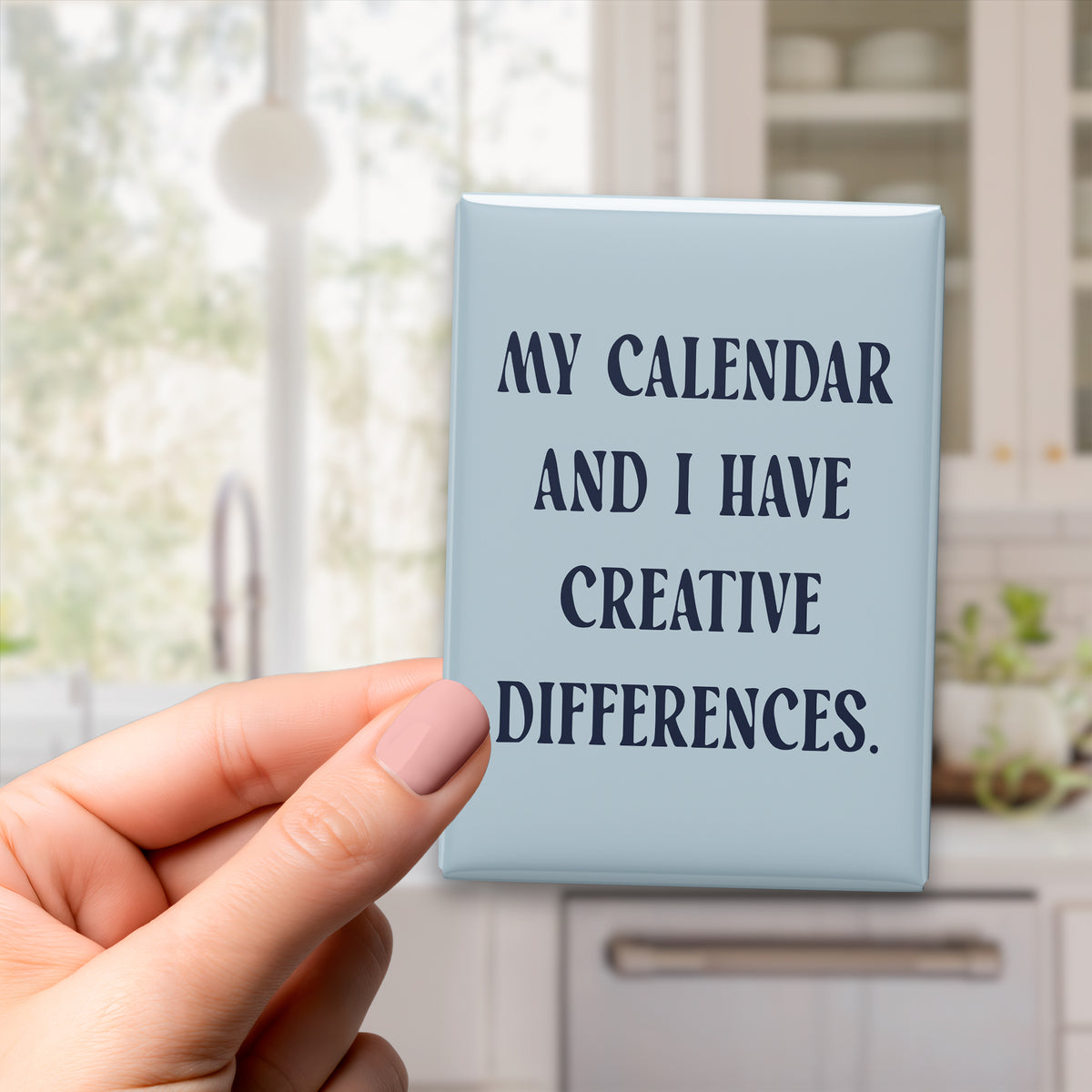 Hand holding the 2.5 × 3.5-inch “My calendar and I have creative differences.” magnet in a kitchen