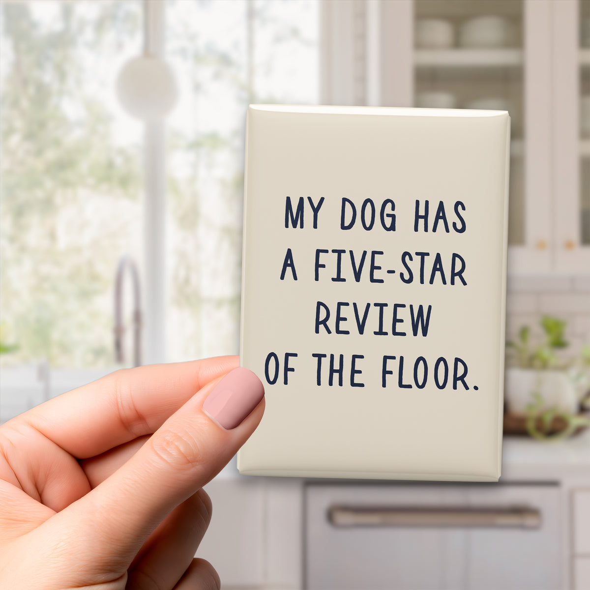 Hand holding the 2.5 × 3.5-inch “My dog has a five-star review of the floor.” magnet in a kitchen