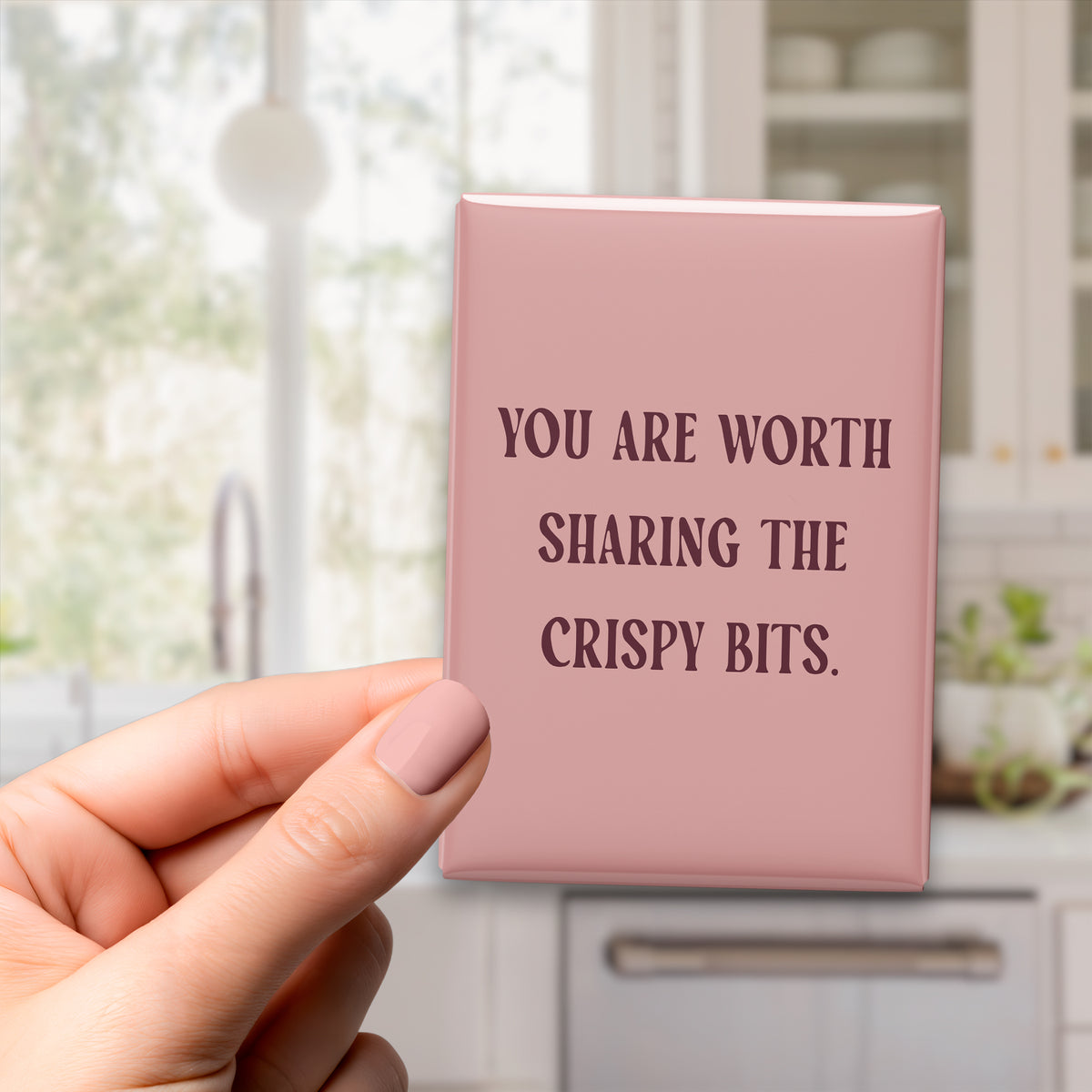 Hand holding the 2.5 × 3.5-inch “You are worth sharing the crispy bits.” magnet in a kitchen