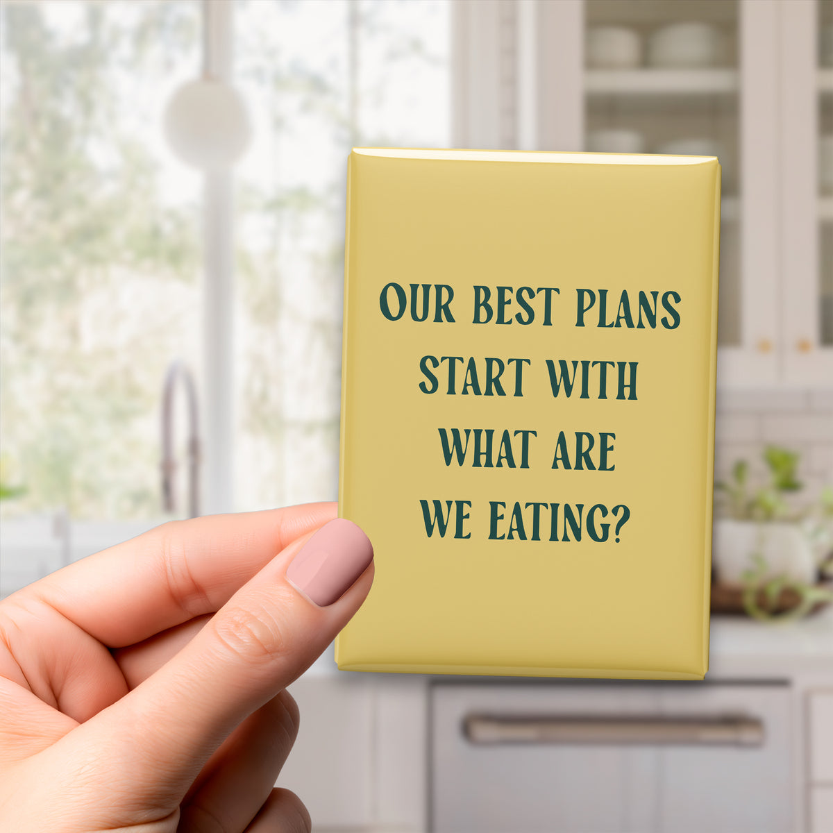 Hand holding the 2.5 × 3.5-inch “Our best plans start with what are we eating?” magnet in a kitchen