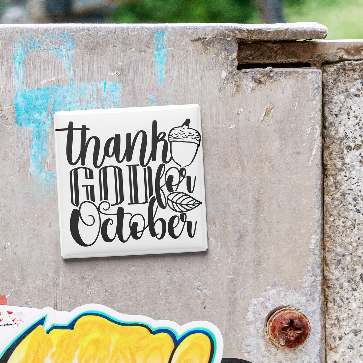 a white square magnet with black text that says "Thank GOD October" attached to a concrete wall.