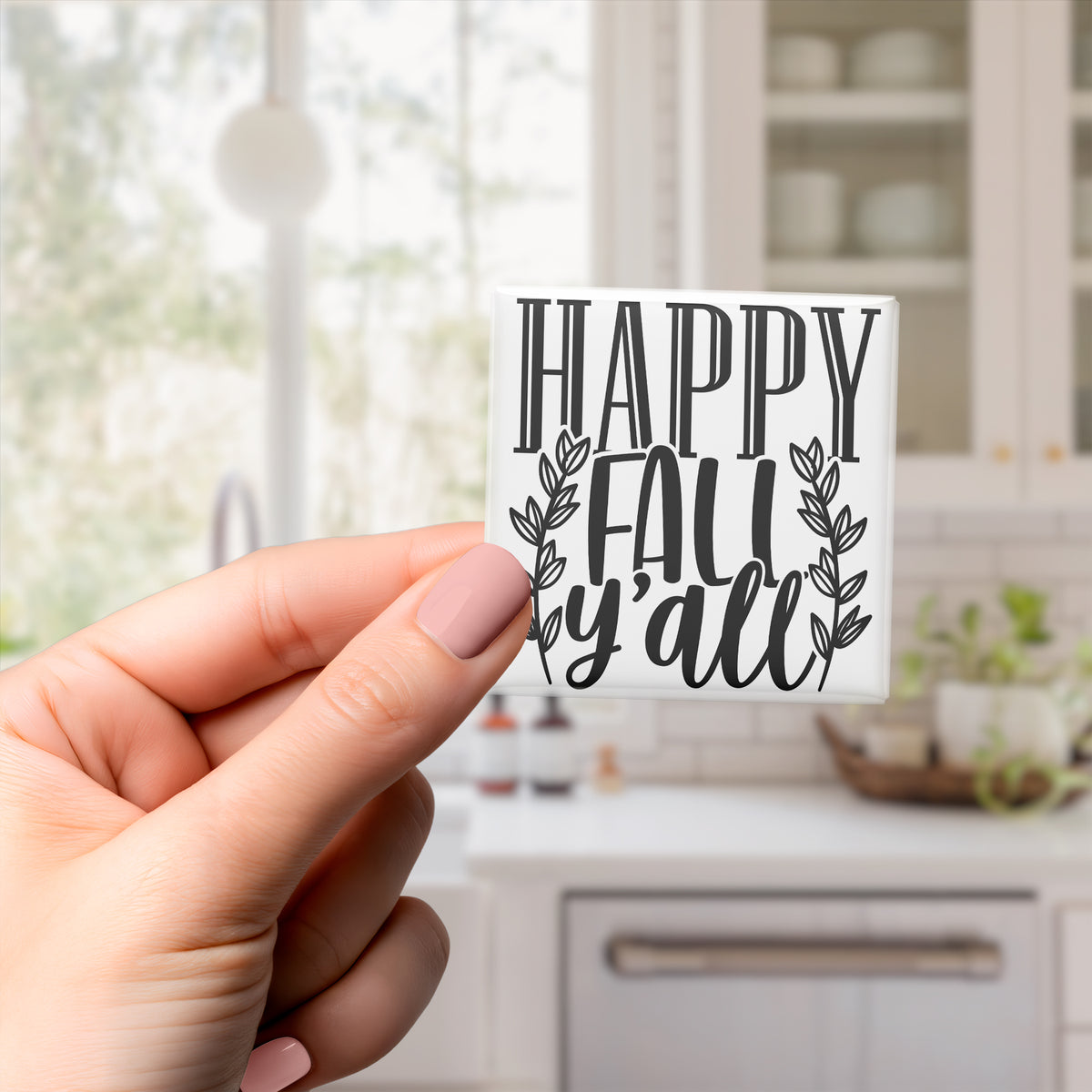 A hand holding a sticker with the text "HAPPY FALL Y'ALL" against a blurred background of a kitchen.