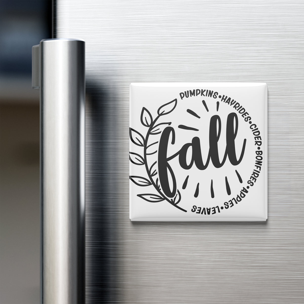 A refrigerator door with a decorative magnet that says "fall" surrounded by a leaf and a sunburst design.