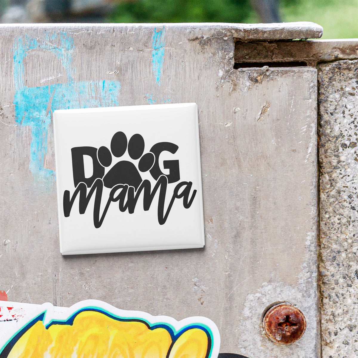 A white square magnet with the word "dog mama" written on it is attached to a weathered concrete surface.