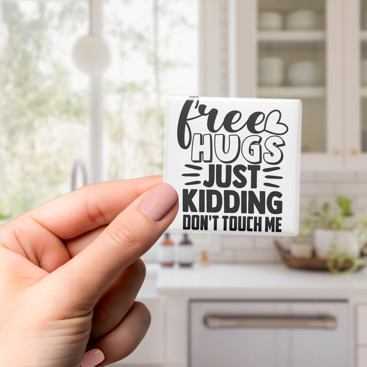 A hand holding a sticker with the text "Free Hugs Just Kidding Don't Touch Me" in a kitchen setting.