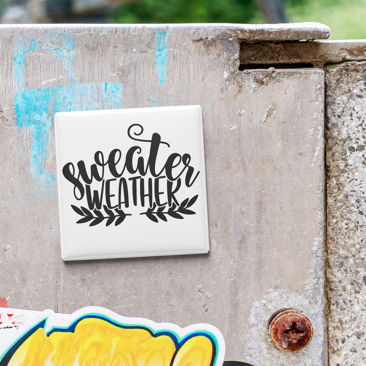 A white square magnet with the text "sweater weather" written on it, attached to a weathered concrete surface.