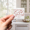 A hand holding a small white square with a heart-shaped design and the text "Be Kind to Yourself" written on it.