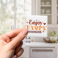 A hand holding a square tile with the text "Enjoy Every moment" on it, with a blurred background of a kitchen counter and cabinets.