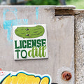 A sticker with a cartoon pickle and the text "License to dill" is attached to a concrete surface.