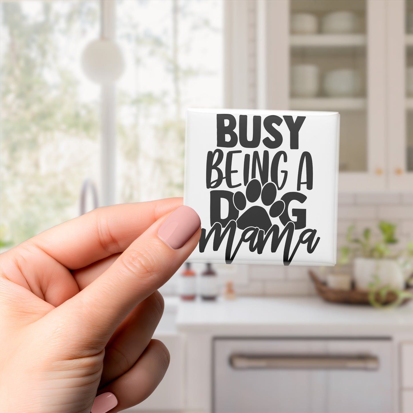 A hand holding a small white sticker with the text "BUSY BEING A DOG MAMA" printed on it.