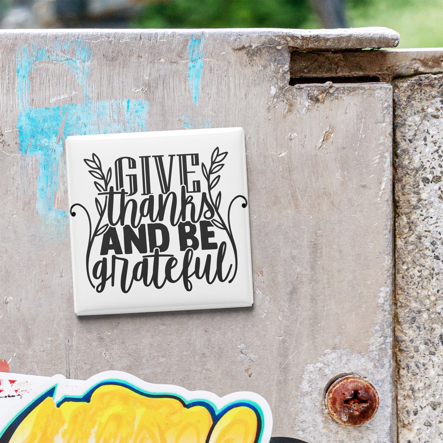 a white tile with the text "GIVE thanks AND BEgrateful" written on it, which is attached to a concrete wall.