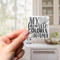 A hand holding a sticker with the text "MY FAVORITE COLORIS AUTUMN" against a blurred background of a kitchen.