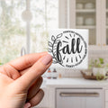 A hand holding a white sticker with the word "fall" written on it, with a blurred background of a kitchen counter and cabinets.