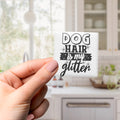A hand holding a small white object with a black text design that says "DOG HAIR is my glitter" against a blurred background of a kitchen counter and cabinets.