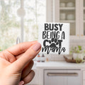 A hand holding a small white object with the text "BUSY BEING A COT MAMA" printed on it.