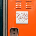 a heart-shaped sticker with the text "Be Kind to Yourself" on an orange surface.