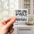 A hand holding a sticker with the text "I LIKE LONG WALKS AWAY FROM PEOPLE" against a blurred background of a kitchen.