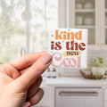 A hand holding a small square magnet with the text "Kind is the new cool" written on it.