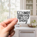 A hand holding a sticker with the text "100% NOT Leaving THE HOUSE TODAY" against a blurred background of a kitchen.