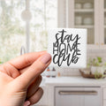 A hand holding a small square magnet with the text "Stay Home Club" written on it.
