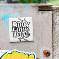 a white square magnet with the text "Enjoy the little things" written on it, placed on a concrete surface.