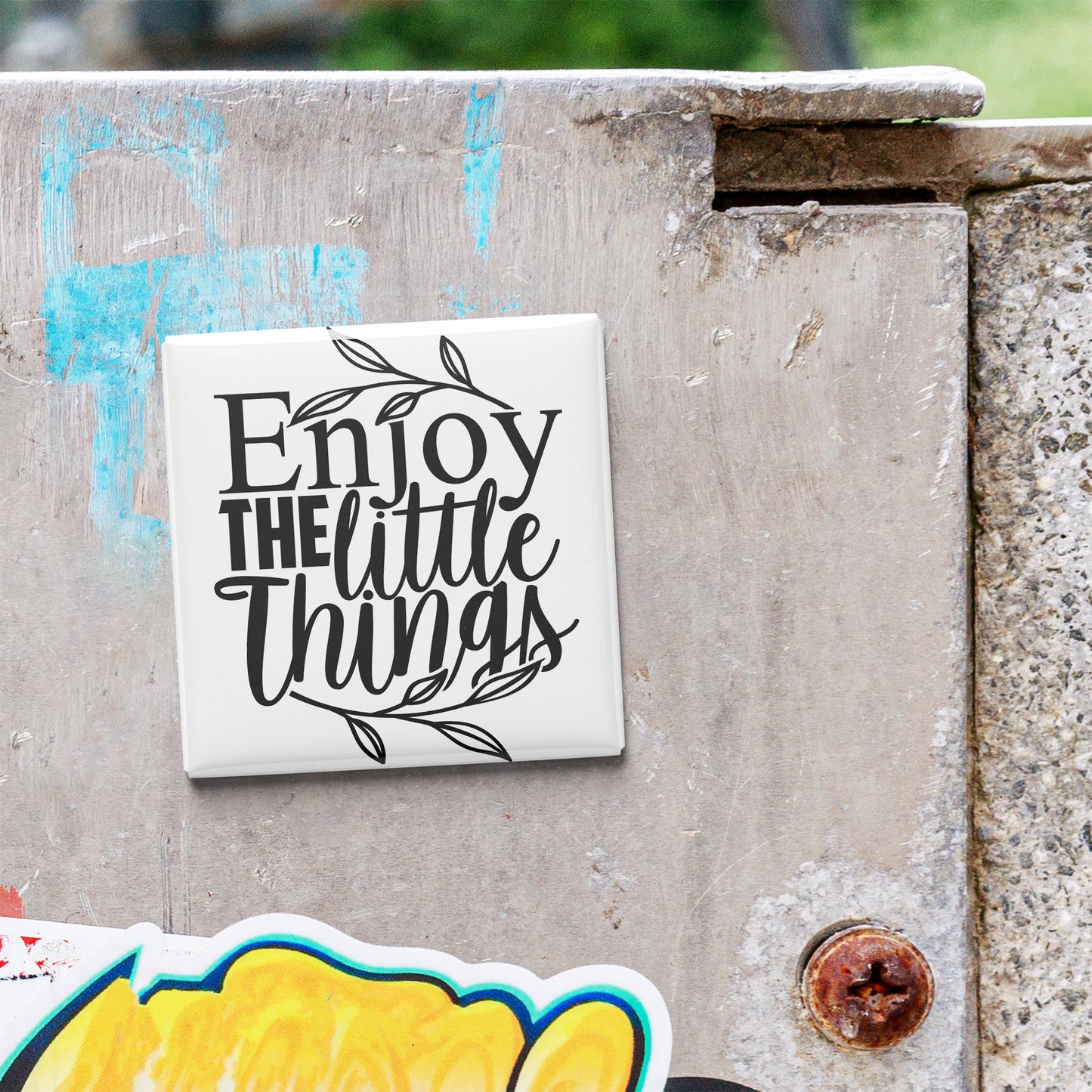 a white square magnet with the text "Enjoy the little things" written on it, placed on a concrete surface.