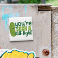 A sticker with a cartoon pickle and the text "you're such a dull light" is attached to a concrete surface.
