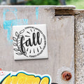 A white tile with a black and white graphic design featuring the word "fall" surrounded by a decorative leaf and the phrase "Pumpkins-Havrils-Cider-Bonfires" in a stylized font.