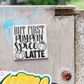 A white sign with black text that says "BUT FIRST PUMPKIN SPICE LATTE" is attached to a concrete wall.