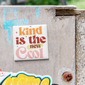 a sticker with the text "kind is the new cool" on a concrete wall.