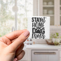 A hand holding a small white object with the text "STAY AT HOME DOG MOM" printed on it.