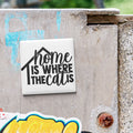 a white square magnet with the text "home is where the cat is" written on it, attached to a weathered concrete surface.
