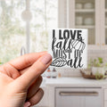 A hand holding a small white sticker with black text that says "I LOVE fall MOST OF all".
