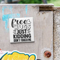 A sticker with the text "Free Hugs Just Kidding Don't Touch Me" is attached to a weathered concrete surface.