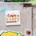 A white square magnet with the text "Enjoy Every moment" is attached to a weathered concrete wall.
