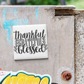 A white square magnet with the text "Thankful, Grateful, Blessed" written on it, attached to a weathered concrete surface.