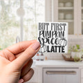 A hand holding a sticker with the text "BUT FIRST PUMPKIN SPICE LATTE" against a blurred background of a kitchen counter and cabinets.