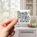 A hand holding a small white sticker with black text that says "Thank GOD October".
